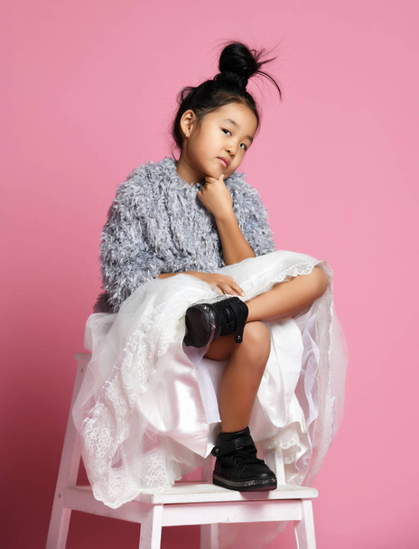Young asian girl kid in long white skirt, grey fluffy sweater and black shoes slegs crossed and think something over - Fotografie, Obrázek