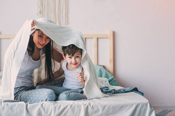 happy mother and son playing and hiding under blanket in bed in cozy weekend morning. Casual lifestyle with real family - Foto, immagini