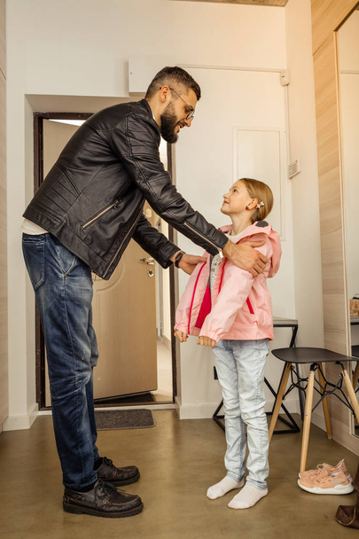 Handsome dark-haired bearded man in eyewear putting a coat on his daughter - Foto, afbeelding