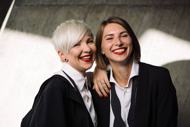 portrait of  two laughing women in classic suits posing against geometrical concrete wall outdoors  - Фото, зображення