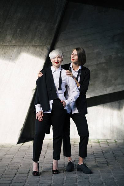 full length shot of two women in classic suits posing against geometrical concrete wall outdoors  - Zdjęcie, obraz