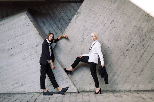 full length shot of two women in classic suits posing against geometrical concrete wall outdoors  - Fotó, kép