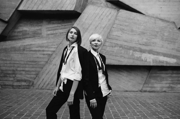 two women in classic suits posing against geometrical concrete wall outdoors  - Fotó, kép
