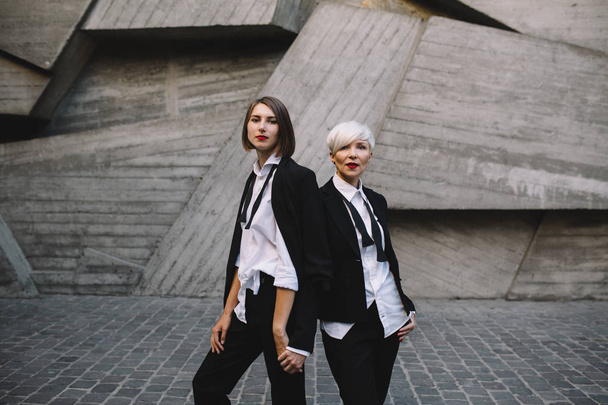 two women in classic suits posing against geometrical concrete wall outdoors  - Фото, зображення