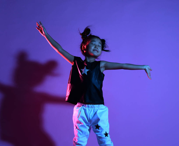 Asian kid girl in shirt and pants with stars and white sneakers dances with closed eyes and jumps - Fotografie, Obrázek