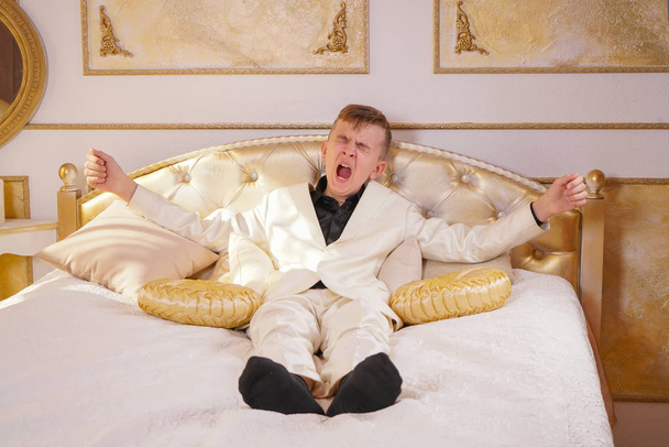 young teen guy wearing white business suit and lying on the bed in his Golden room, stretching and yawning - Photo, Image