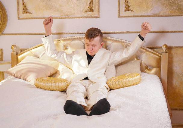 Молодой подросток в белом деловом костюме лежит на кровати в своей Золотой комнате, растягиваясь и зевая
 - Фото, изображение