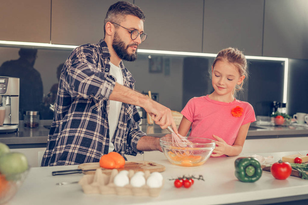 Bearded dark-haired man in eyewear beating up eggs in the plate - Photo, image