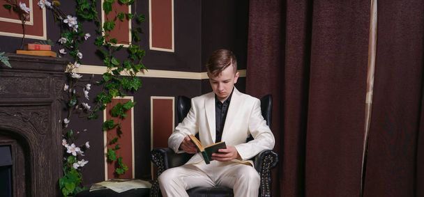znudzony student Nastolatki facet w białym garniturze, siedząc w fotelu z książką przy kominku - Zdjęcie, obraz