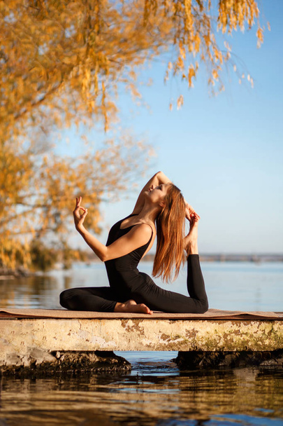 Young woman practicing yoga exercise at quiet pier in autumn park. - Фото, изображение