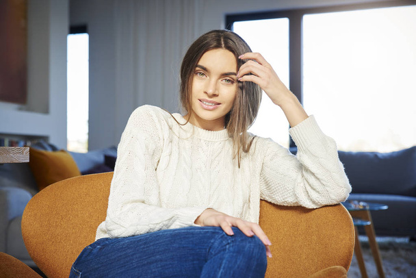 Portrait shot of smiling young woman wearing casual clothes while relaxing in armchair at home.  - Foto, Bild