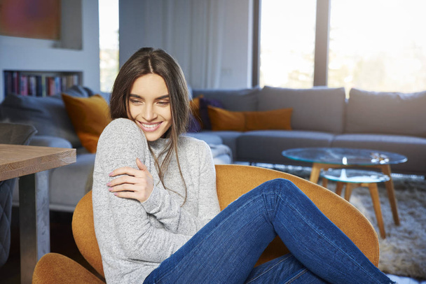 Portrait shot of beautiful young woman wearing casual clothes and relaxing in armchair while daydreaming at home. - Foto, Imagem