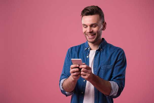 Young handsome modern casual man with smart phone in hands is plating in online game isolated on pink background - Foto, imagen