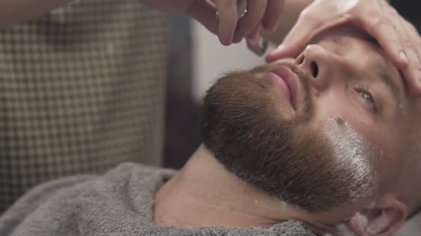 Barber shaving bearded man with straight razor in male salon. Male skin care - Filmati, video
