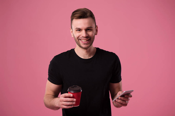 Young handsome modern casual man with smart phone and coffee looks on camera isolated on pink background - Φωτογραφία, εικόνα