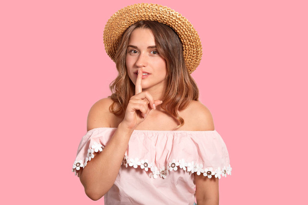 Shh, dont tell this to anybody. Secret woman keeps fore finger over mouth, tells confidential information, wears straw hat and fashionable blouse, poses against pink background. Hush concept - Photo, Image