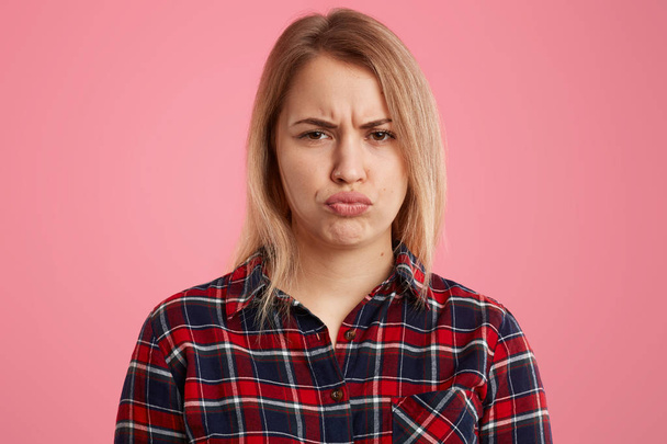 Indoor shot of displeased blonde woman pouts lips and frowns face, doesnt like something, has sullen facial expression, wears checkered shirt, isolated over pink background, makes dissatisfied grimace - 写真・画像