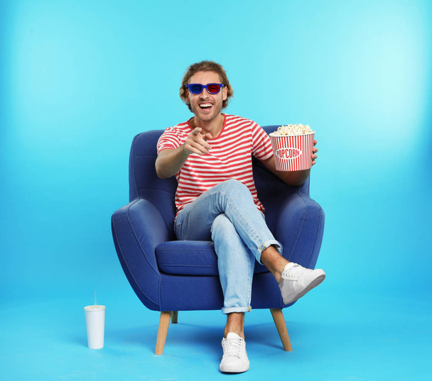 Man with 3D glasses, popcorn and beverage sitting in armchair during cinema show on color background - Foto, imagen