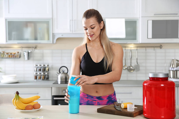Young woman preparing protein shake at table in kitchen - Foto, afbeelding