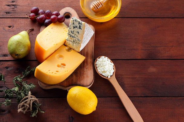 Hard cheese, blue cheese and cottage cheese on wooden board. Different types of cheese and honey on wooden background. Dairy products, pear, lemon and grapes. Top view - Foto, Imagen