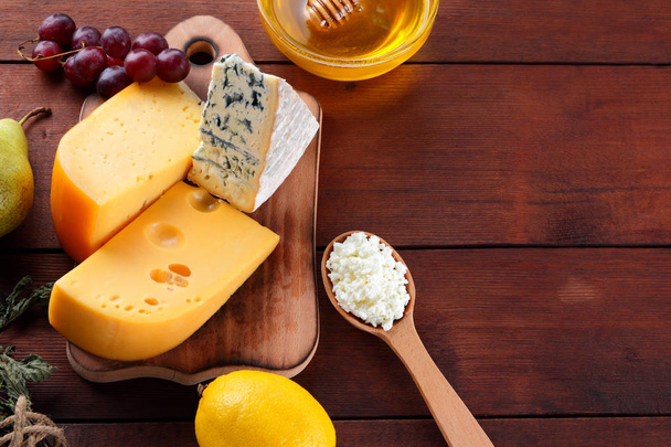 Hard cheese, blue cheese and cottage cheese on wooden board. Different types of cheese and honey on wooden background. Dairy products, pear, lemon and grapes. Top view - Foto, Imagen