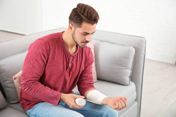 Young man applying bandage on injured arm at home. First aid - Zdjęcie, obraz