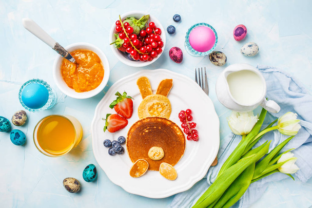 Великодній кролик з млинцями з ягодами. Сніданок на Великдень. Кольорові яйця, молоко, сік і джем. Синій фон, вид зверху
. - Фото, зображення