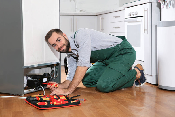Male technician in uniform repairing refrigerator indoors - Foto, afbeelding
