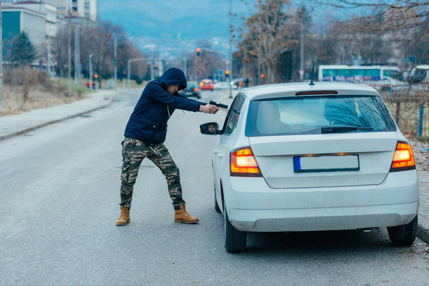 Гангстер с пистолетом пытается ограбить парня, который сидит в своей машине
. - Фото, изображение