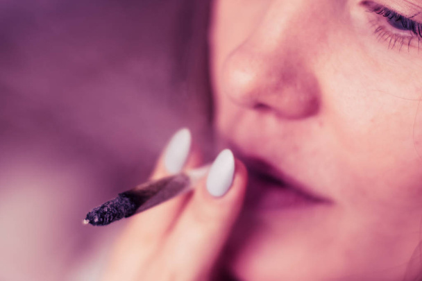 joint in woman hand. A woman smokes cannabis weed, a . Smoke on a black background. Concepts of medical marijuana use and legalization of the cannabis. On a black background Pink tinting - Фото, зображення
