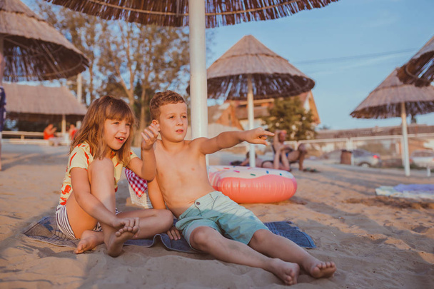Onnellisia positiivisia lapsia istuu pyyhkeellä ja pitää hauskaa hiekkarannalla
 - Valokuva, kuva