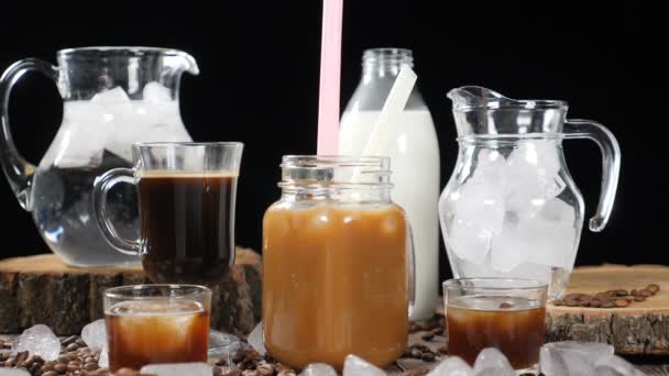 Dlouhý záběr sady lahví a sklenic s mlékem, voda, černá káva a karamelový nápoj. Kostky ledu všude kolem. Ženská ruka uvedení slámy ve sklenici ve zpomaleném filmu. HD - Záběry, video