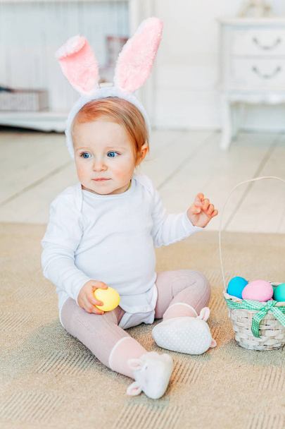 Little child girl wearing bunny ears on Easter day. Girl holding basket with painted eggs sitting on floor at home, having fun on Easter egg hunt. Happy Easter holiday celebration spring concept - Photo, Image