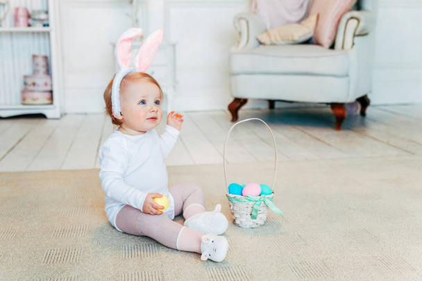 Маленькая девочка с кроличьими ушами на Пасху. Девушка держит корзину с покрашенными яйцами, сидя дома на полу, развлекаясь в пасхальной охоте за яйцами. С праздником Пасхи!
 - Фото, изображение