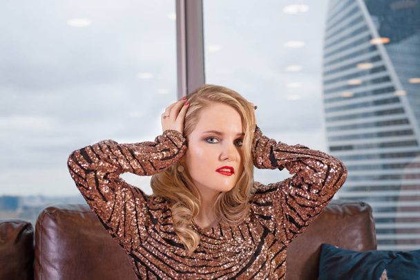 Une belle jeune fille blonde dans une robe à la mode posant sur un canapé en cuir sur le fond d'une fenêtre panoramique donnant sur les gratte-ciel
 - Photo, image