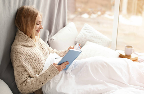 Beautiful young woman in knitted sweater sitting and reading book near window at home. Winter atmosphere - Fotoğraf, Görsel