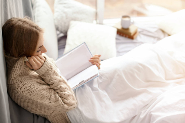 Gyönyörű fiatal nő a kötött pulóvert, ülő- és olvasási könyv közelében ablak otthon. Téli hangulatot - Fotó, kép