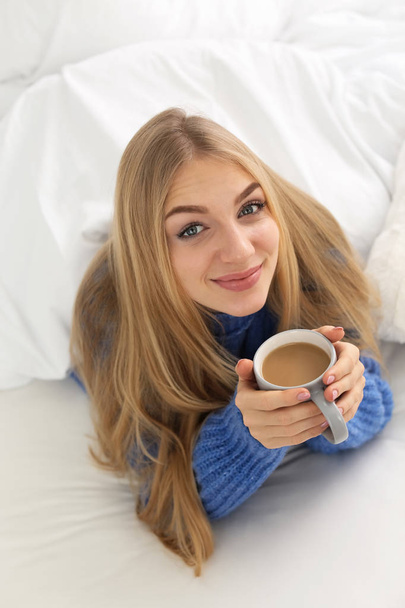 Beautiful young woman with cup lying in bed at home. Winter atmosphere - Zdjęcie, obraz