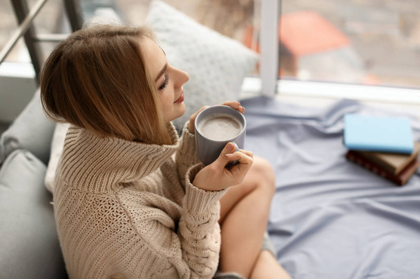 Beautiful young woman in knitted sweater sitting with cup of coffee at home. Winter atmosphere - Foto, afbeelding