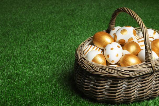 Wicker basket of traditional Easter eggs decorated with golden paint on green lawn, space for text - Φωτογραφία, εικόνα