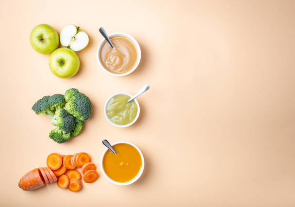 Белые миски со здоровым натуральным детским питанием на пастельном фоне с местом для текста. Свежие фрукты и овощи пюре, мясорубка, вид сверху, концепция. Детская еда из моркови, брокколи, яблок
 - Фото, изображение
