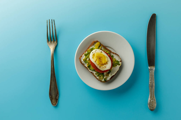 Healthy green vegetarian toast with avocado, tomato, egg. healthy eating, vegan diet concept. food on a plate on a blue background with a fork and knife - Photo, image