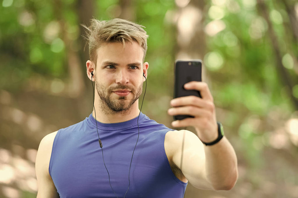 Sport gadget concept. Athlete mobile phone set up playlist before runnig. Man athlete busy face setting up smartphone app, nature background. Sportsman training with pedometer and earphones - Photo, Image