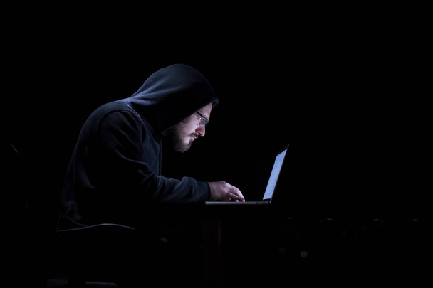 Jeune hacker talentueux utilisant un ordinateur portable tout en travaillant dans un bureau sombre avec de grandes lumières de la ville en arrière-plan la nuit - Photo, image