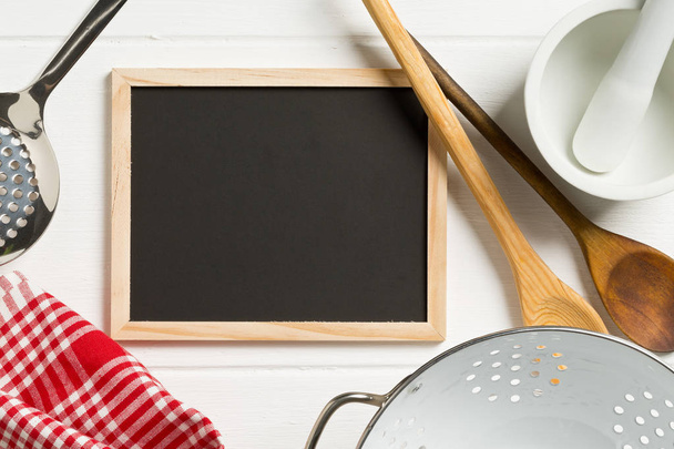 Blank, empty, black chalkboard with cooking utensils and red checkered dish towel flat lay from above on white wooden table with copy space - Fotografie, Obrázek