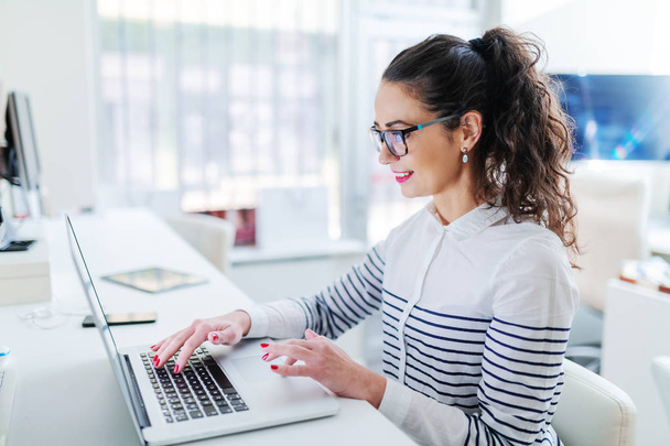 Zijaanzicht van hard werkende vrouwelijke architect laptop gebruikt en zit in het kantoor. Witte kleur domineert. - Foto, afbeelding