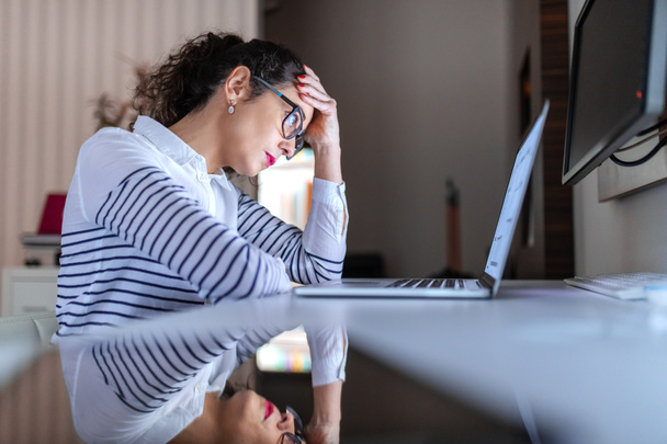 Visão lateral da preocupação trabalhadora secretária feminina segurando a mão na cabeça e olhando para o laptop enquanto se inclina na mesa
. - Foto, Imagem