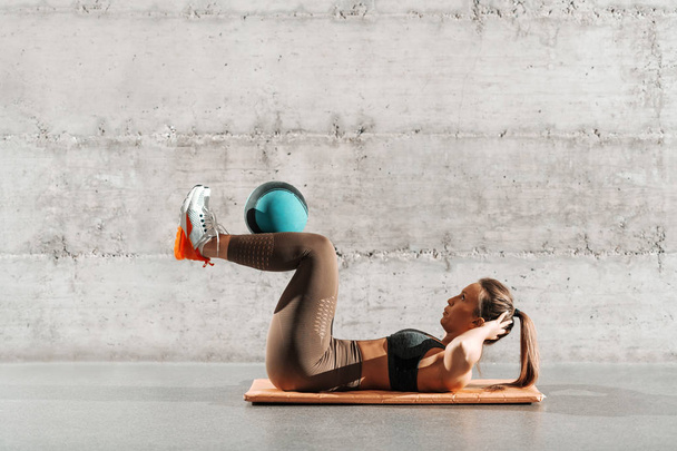 Side view of muscular powerful woman with ponytail and in sportswear doing abs with ball on the mat in front of gray wall. - Photo, Image