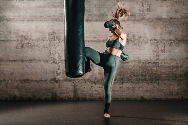 Dedicated strong brunette with ponytail, in sportswear, bare foot and with boxing gloves kicking sack in gym. - Photo, Image