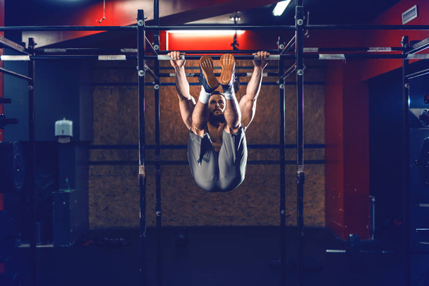 Shirtless Caucasian bearded bodybuilder doing chin ups with legs up. Gym interior, night work out concept. - Fotoğraf, Görsel